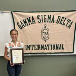 Haylee Ferguson holding her Outstanding Senior Award standing in front of the Gamma Sigma Delta International banner